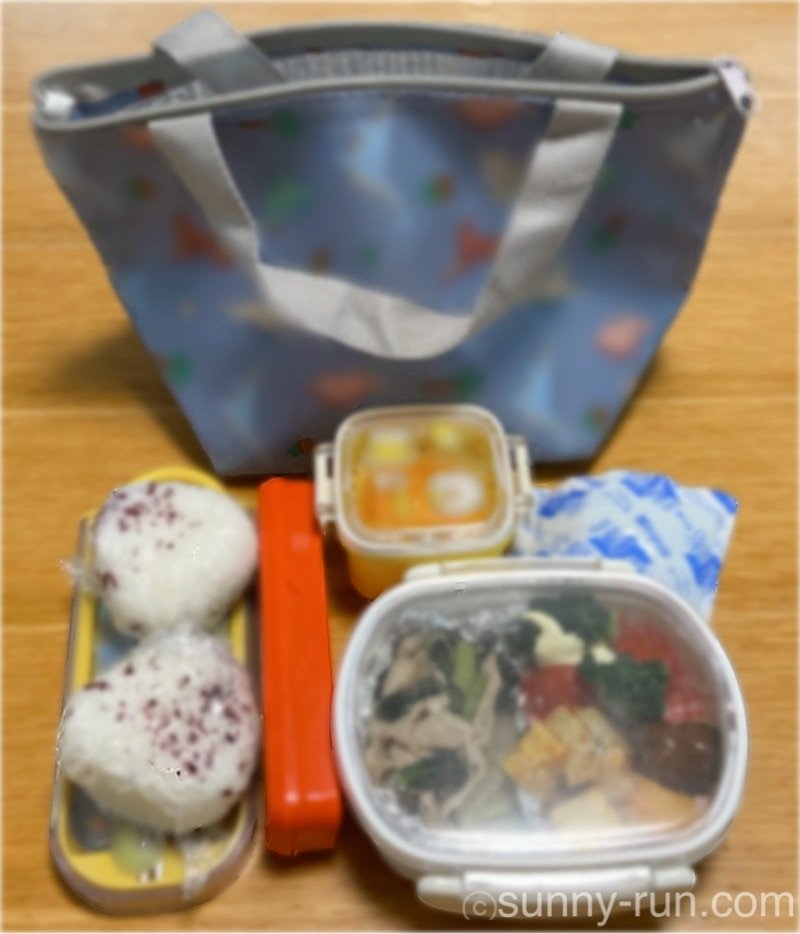 お弁当バッグの中身(お弁当箱、デザート入れ、おにぎり2つ、カトラリーケース、おしぼり入れ、保冷剤)を机の上に並べた様子